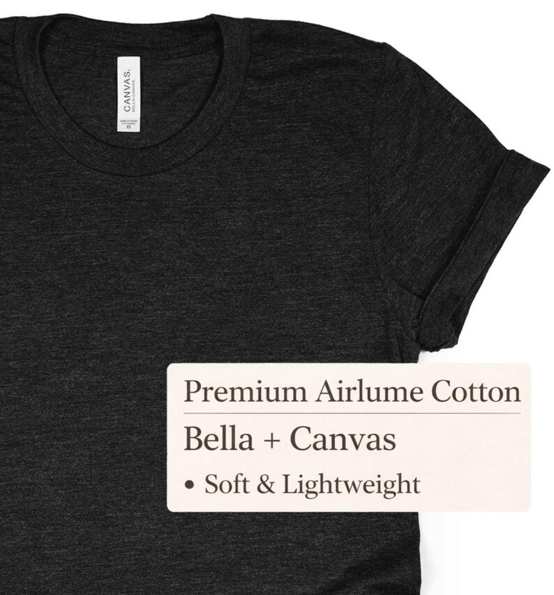 K&ouml;nnte beinhalten: Dunkelgraues T-Shirt mit hochgekrempelten &Auml;rmeln. Das Shirt hat einen Rundhalsausschnitt und besteht aus einem weichen, leichten Material. Das Etikett lautet "Premium Airlume Cotton" und "Bella + Canvas".
