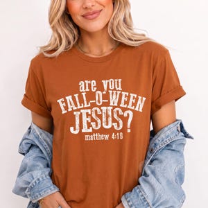 May include: A burnt orange short-sleeve t-shirt featuring white distressed text that reads are you FALL-O-WEEN JESUS? matthew 4:19. The shirt has a crew neckline and is worn with a light blue denim jacket draped over the shoulders.