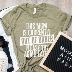 K&ouml;nnte beinhalten: Ein olivgr&uuml;nes T-Shirt mit dem Text "THIS MOM IS CURRENTLY OUT OF ORDER PLEASE TRY AGAIN LATE." Neben dem Shirt befindet sich eine schwarze Buchstaben-Tafel mit der Aufschrift "MOMMIN' AIN'T EASY." Auch Sonnenbrillen, Schuhe und andere Shirts sind zu sehen.