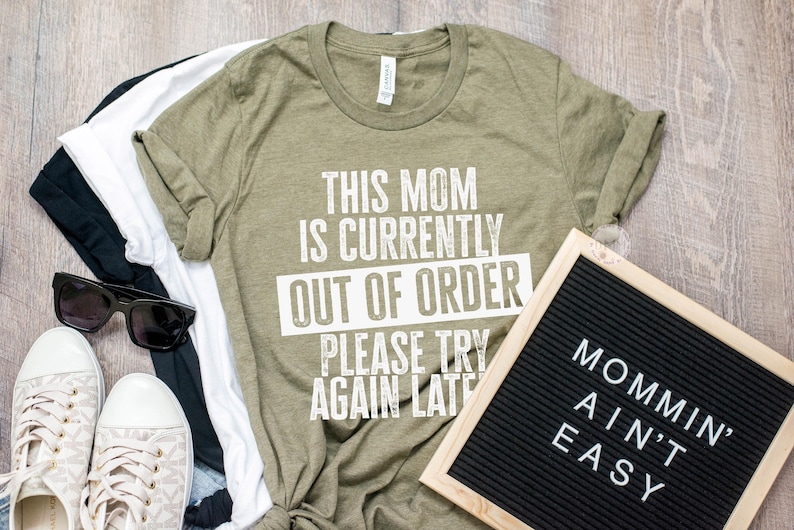 K&ouml;nnte beinhalten: Ein olivgr&uuml;nes T-Shirt mit dem Text "THIS MOM IS CURRENTLY OUT OF ORDER PLEASE TRY AGAIN LATE." Neben dem Shirt befindet sich eine schwarze Buchstaben-Tafel mit der Aufschrift "MOMMIN' AIN'T EASY." Auch Sonnenbrillen, Schuhe und andere Shirts sind zu sehen.