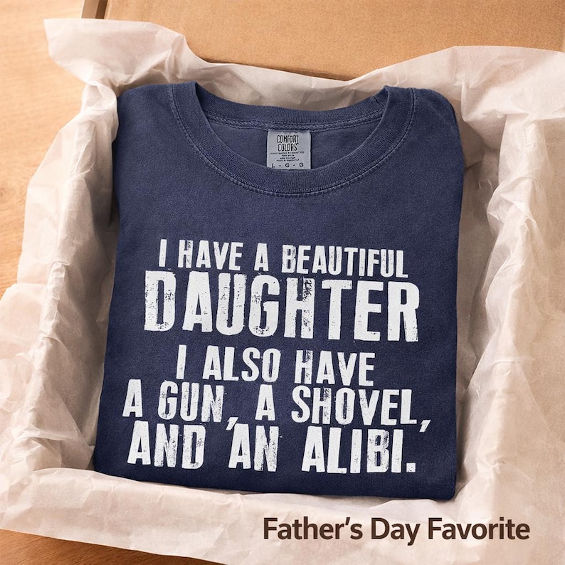 May include: A navy blue crew neck t-shirt with white distressed text that reads I HAVE A BEAUTIFUL DAUGHTER I ALSO HAVE A GUN, A SHOVEL, AND AN ALIBI. The shirt features a Comfort Colors brand label at the inner neckline indicating a size large.