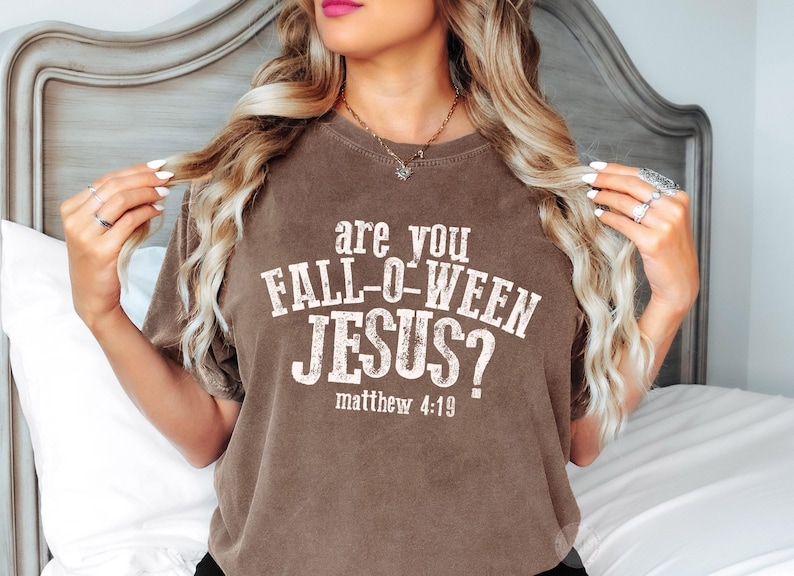 Puede incluir: Una camiseta marr&oacute;n de manga corta con el texto blanco desgastado "are you FALL-O-WEEN JESUS?" seguido de "matthew 4:19" impreso en la parte delantera. La prenda tiene cuello redondo y un corte holgado con una est&eacute;tica vintage.