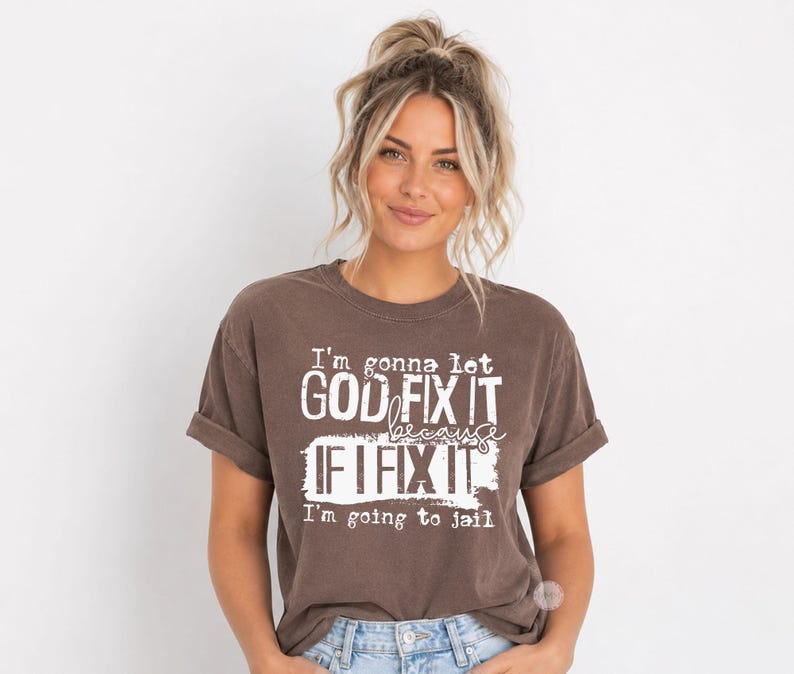 May include: A brown short-sleeved t-shirt with a crew neckline and rolled sleeves. The front features white distressed-style text that reads: I'm gonna let GOD FIX IT because IF I FIX IT I'm going to jail. The shirt has a relaxed fit.