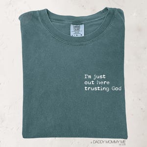 K&ouml;nnte beinhalten: Ein blau-fichtenfarbenes T-Shirt mit Rundhalsausschnitt und einem Comfort Colors Markenetikett am Innenkragen. Auf der Vorderseite befindet sich der wei&szlig;e Schriftzug "I'm just out here trusting God" in einer abgenutzten, strukturierten Schrift.