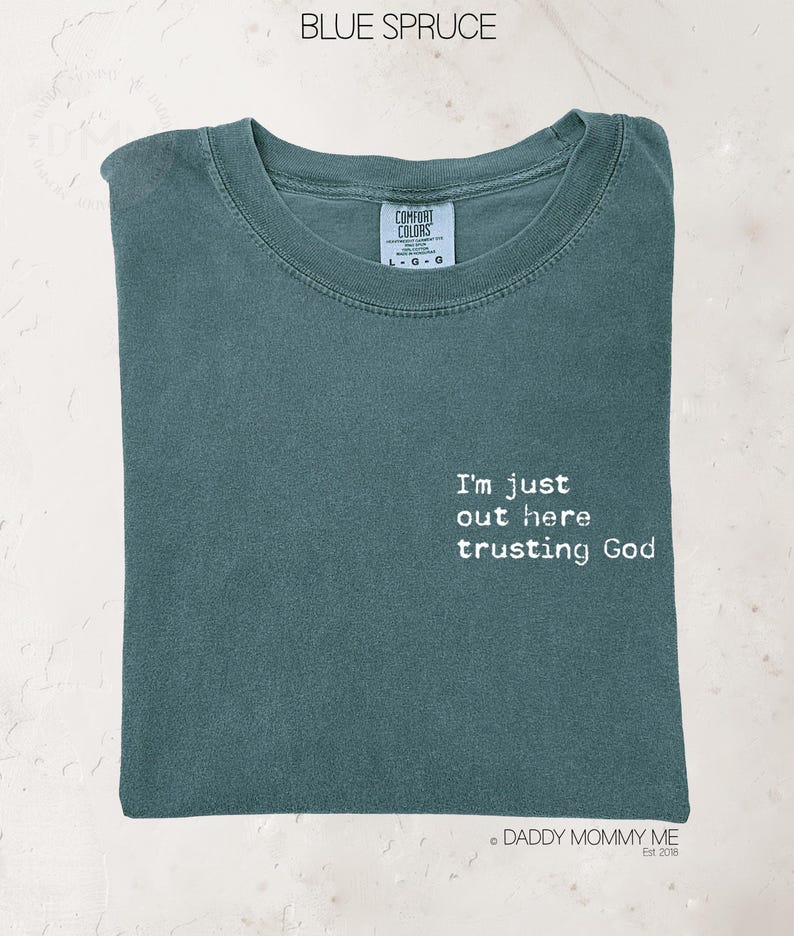 K&ouml;nnte beinhalten: Ein blau-fichtenfarbenes T-Shirt mit Rundhalsausschnitt und einem Comfort Colors Markenetikett am Innenkragen. Auf der Vorderseite befindet sich der wei&szlig;e Schriftzug "I'm just out here trusting God" in einer abgenutzten, strukturierten Schrift.