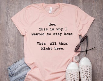 May include: A light pink t-shirt with the text "See. This is why I wanted to stay home. This. All this. Right here." Black sunglasses and white sneakers are next to the shirt.