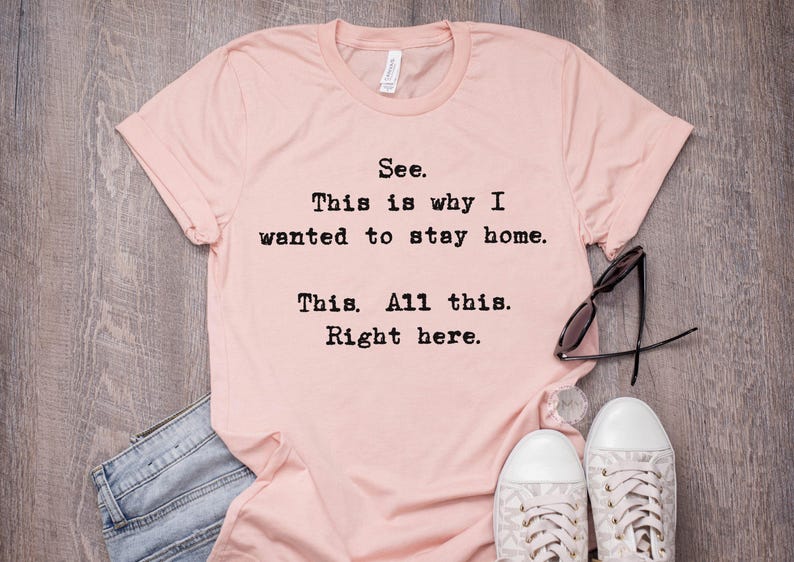 May include: A light pink t-shirt with the text "See. This is why I wanted to stay home. This. All this. Right here." Black sunglasses and white sneakers are next to the shirt.