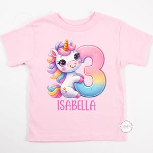 May include: A light pink t-shirt featuring a colorful unicorn design with a rainbow mane and tail, and the number 3. The name "ISABELLA" is printed below the unicorn in pink letters. This shirt is perfect for a child's birthday.