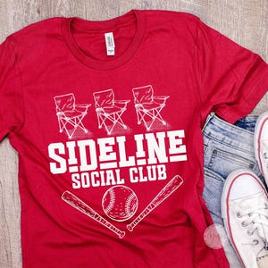 Op de afbeelding: Rood t-shirt met de witte tekst "SIDELINE SOCIAL CLUB", met afbeeldingen van klapstoelen, een honkbal en knuppels. Het shirt wordt gecombineerd met een blauwe jeans en witte sneakers.