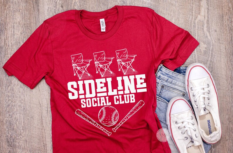 Op de afbeelding: Rood t-shirt met de witte tekst "SIDELINE SOCIAL CLUB", met afbeeldingen van klapstoelen, een honkbal en knuppels. Het shirt wordt gecombineerd met een blauwe jeans en witte sneakers.