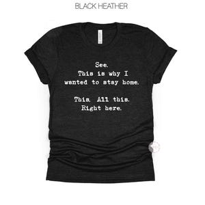 May include: Black heather t-shirt with the text "See. This is why I wanted to stay home. This. All this. Right here." The shirt has a V-neck and short sleeves. The text is in a simple, sans-serif font.
