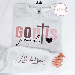 May include: A gray sweatshirt with the text "GOD IS good" in pink and brown letters with a black heart and a cross. The text "All the times" is written in cursive below the main text.