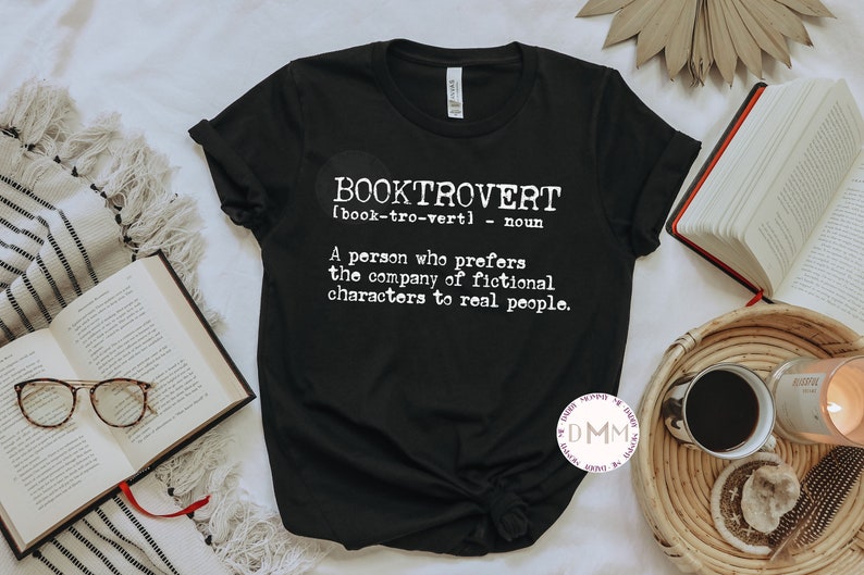 May include: Black t-shirt with the word "BOOKTROVERT" in white, along with a definition. The shirt is styled with open books, eyeglasses, and a coffee cup. The definition reads: "A person who prefers the company of fictional characters to real people."