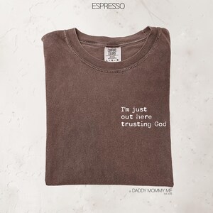 K&ouml;nnte beinhalten: Ein espressofarbenes T-Shirt mit Rundhalsausschnitt und einem Comfort Colors-Etikett. Auf der Vorderseite befindet sich ein wei&szlig;er Schriftzug mit dem Text, I'm just out here trusting God. Das T-Shirt hat kurze &Auml;rmel und eine Standardpassform.