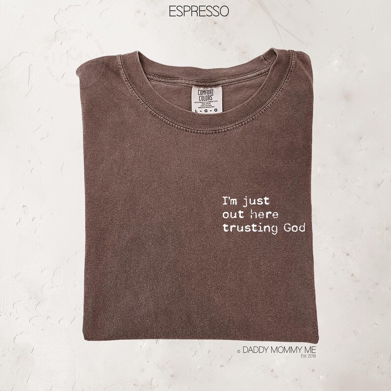 K&ouml;nnte beinhalten: Ein espressofarbenes T-Shirt mit Rundhalsausschnitt und einem Comfort Colors-Etikett. Auf der Vorderseite befindet sich ein wei&szlig;er Schriftzug mit dem Text, I'm just out here trusting God. Das T-Shirt hat kurze &Auml;rmel und eine Standardpassform.