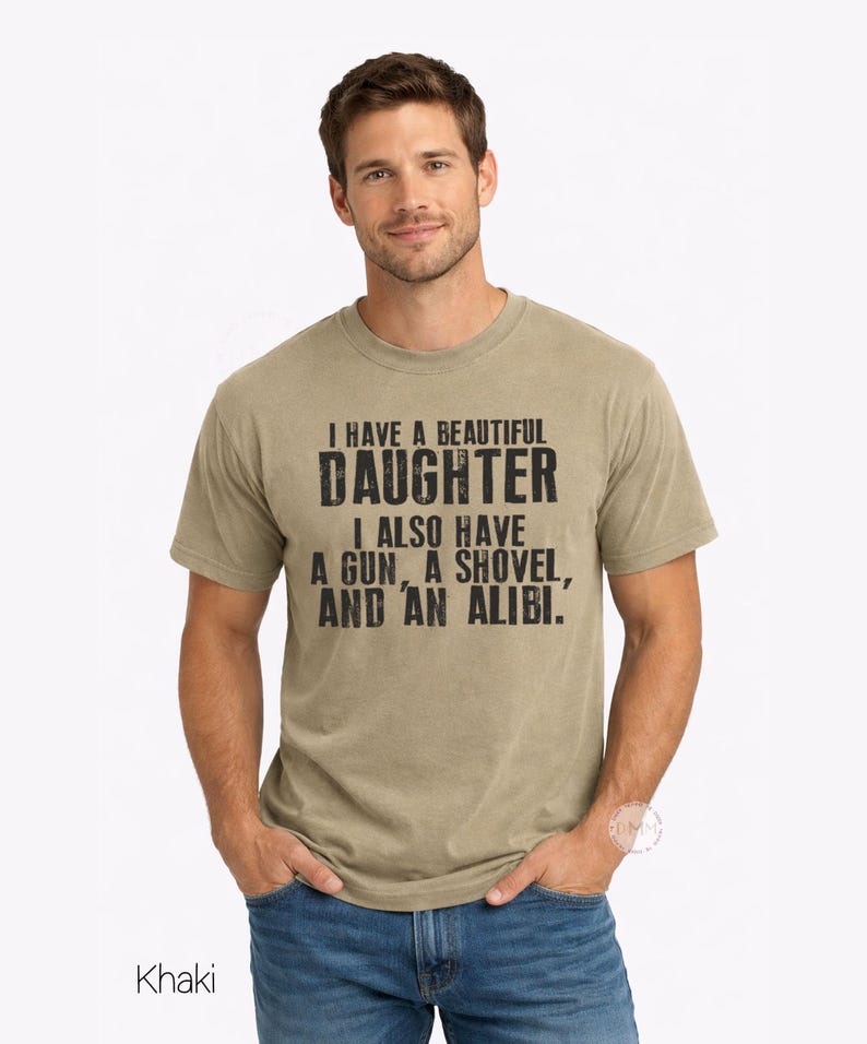 May include: A khaki short-sleeved crew neck t-shirt featuring black distressed text that reads: I HAVE A BEAUTIFUL DAUGHTER I ALSO HAVE A GUN, A SHOVEL, AND AN ALIBI. The shirt is paired with blue denim jeans, creating a casual look for everyday wear.
