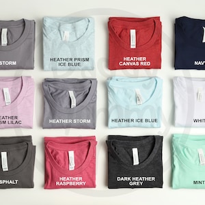 K&ouml;nnte beinhalten: Eine Sammlung gefalteter T-Shirts in verschiedenen Farben, darunter Grau, Blau, Rot und Wei&szlig;. Jedes Shirt hat ein Etikett mit dem Farbnamen, wie "Storm", "Navy" und "Mint". Die Shirts sind ordentlich angeordnet und zeigen eine Reihe von Farbt&ouml;nen.