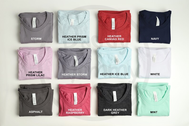 K&ouml;nnte beinhalten: Eine Sammlung gefalteter T-Shirts in verschiedenen Farben, darunter Grau, Blau, Rot und Wei&szlig;. Jedes Shirt hat ein Etikett mit dem Farbnamen, wie "Storm", "Navy" und "Mint". Die Shirts sind ordentlich angeordnet und zeigen eine Reihe von Farbt&ouml;nen.