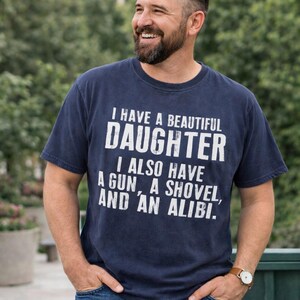 May include: A navy blue short-sleeved t-shirt featuring white distressed text that reads: I HAVE A BEAUTIFUL DAUGHTER, I ALSO HAVE A GUN, A SHOVEL, AND AN ALIBI. The shirt has a crew neckline and a standard fit, displaying the printed graphic clearly.
