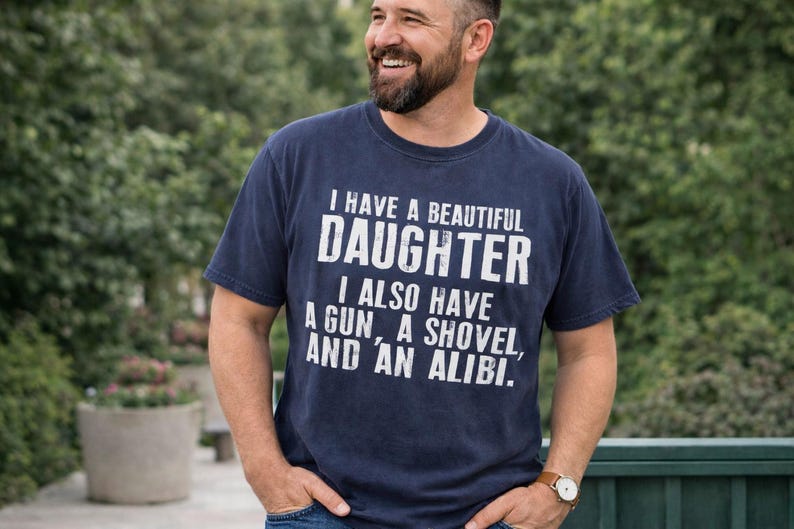May include: A navy blue short-sleeved t-shirt featuring white distressed text that reads: I HAVE A BEAUTIFUL DAUGHTER, I ALSO HAVE A GUN, A SHOVEL, AND AN ALIBI. The shirt has a crew neckline and a standard fit, displaying the printed graphic clearly.