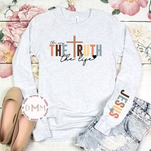 May include: A light gray long-sleeve shirt with the words "the way, the truth, the life" and a cross graphic. The sleeve features the word "JESUS" in colorful letters. The shirt is paired with denim shorts and beige flats.