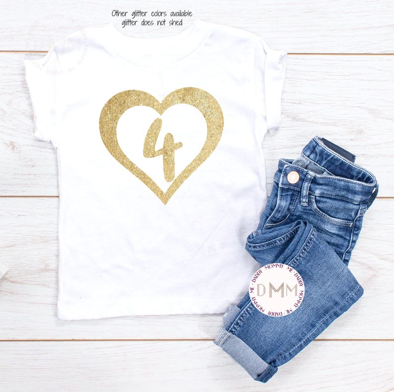 May include: White t-shirt with a gold glitter heart with the number 4 inside. The shirt is laying on a white wooden surface with a pair of blue jeans folded next to it.