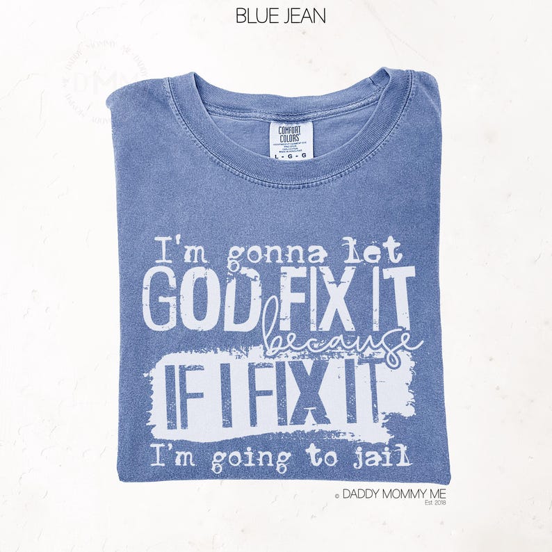 May include: A blue jean colored t-shirt with white screen-printed text that reads: I'm gonna let GOD FIX IT because IF I FIX IT I'm going to jail. The shirt features a crew neckline and a Comfort Colors brand label at the inner collar area.