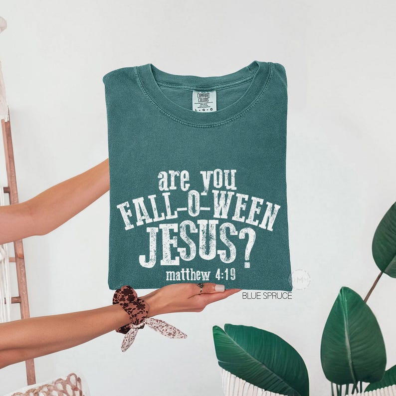 Puede incluir: Una camiseta de manga corta color verde azulado con cuello redondo y texto blanco desgastado que dice "are you FALL-O-WEEN JESUS? matthew 4:19". La prenda tiene una etiqueta de marca visible en el cuello interior y un aspecto de tela suave.