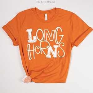 May include: Burnt orange t-shirt with rolled sleeves. The shirt features the white text "LONG HORNS" in a playful, outlined font. The shirt is made of a soft material and is likely a unisex fit.