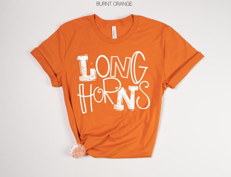 May include: Burnt orange t-shirt with rolled sleeves. The shirt features the white text "LONG HORNS" in a playful, outlined font. The shirt is made of a soft material and is likely a unisex fit.