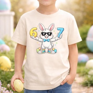 May include: A beige t-shirt featuring a cartoon Easter bunny wearing sunglasses and a bow tie, holding the numbers 6 and 7. The bunny is surrounded by colorful Easter eggs and wearing sneakers. The child is holding a yellow egg.