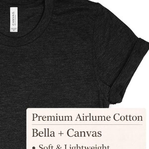 May include: A close-up shot of a dark grey t-shirt with a rolled-up sleeve. The shirt features a crew neck and is made of a soft, lightweight material. A white label with text: "Premium Airlume Cotton" and "Bella + Canvas".