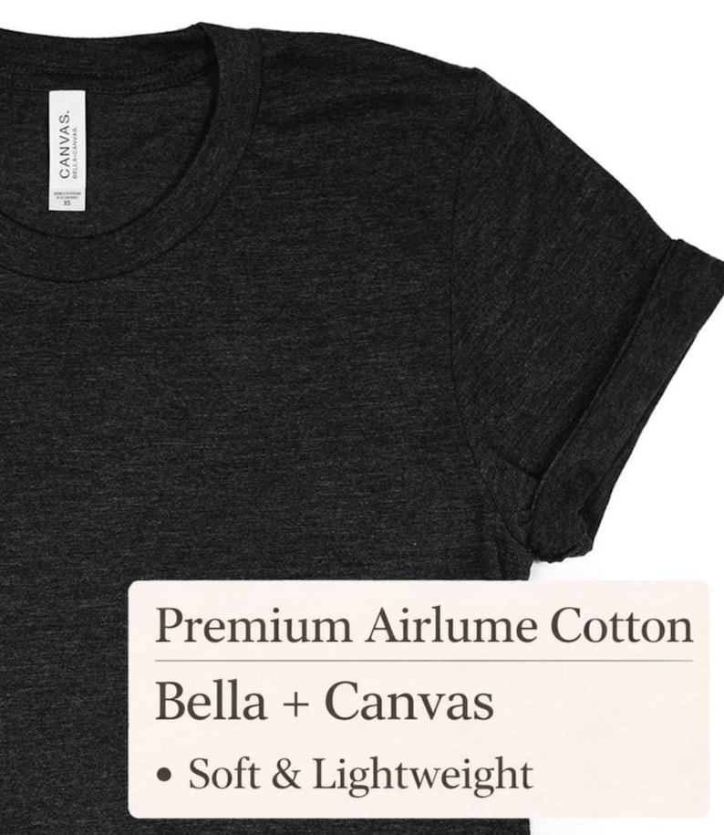 May include: A close-up shot of a dark grey t-shirt with a rolled-up sleeve. The shirt features a crew neck and is made of a soft, lightweight material. A white label with text: "Premium Airlume Cotton" and "Bella + Canvas".