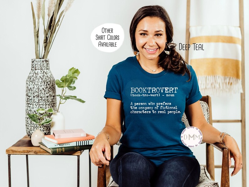 May include: Deep teal t-shirt with the word "BOOKTROVERT" and its definition printed in white. The shirt is a crew neck and has rolled-up sleeves. The text "OTHER SHIRT COLORS AVAILABLE" is in a white circle in the upper left corner.