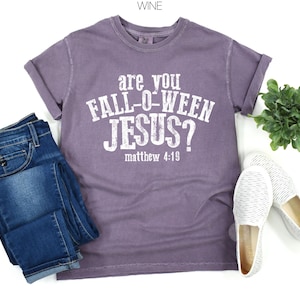 Puede incluir: Una camiseta de manga corta color vino con texto blanco desgastado que dice "are you FALL-O-WEEN JESUS? matthew 4:19". La camiseta se combina con pantalones vaqueros azules y un par de zapatos blancos sin cordones, junto a una peque&ntilde;a planta verde.