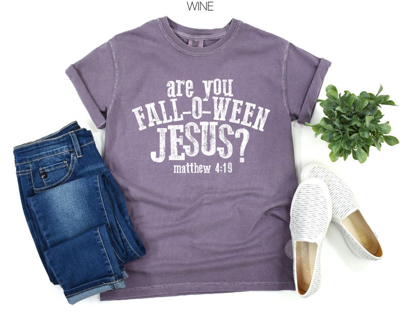 Puede incluir: Una camiseta de manga corta color vino con texto blanco desgastado que dice "are you FALL-O-WEEN JESUS? matthew 4:19". La camiseta se combina con pantalones vaqueros azules y un par de zapatos blancos sin cordones, junto a una peque&ntilde;a planta verde.