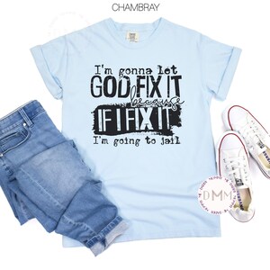 May include: A light blue short-sleeve t-shirt with black text that reads: I'm gonna let GOD FIX IT because IF I FIX IT I'm going to jail. The shirt is paired with blue denim jeans and a pair of white low-top canvas sneakers with white laces.