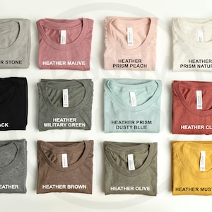 May include: An assortment of folded t-shirts in shades like heather stone, mauve, peach, and natural. Each shirt displays a tag and a label indicating the colour, such as "Black" and "Mustard". The shirts are neatly arranged on a white surface.