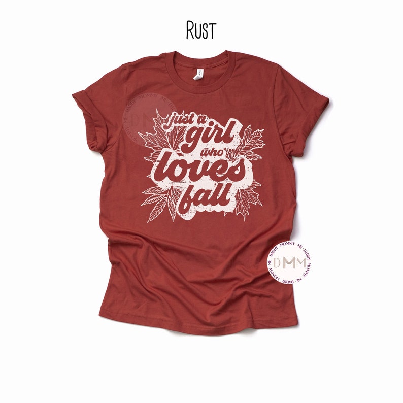 May include: Rust-colored t-shirt with the text "just a girl who loves fall" in a white, distressed font. The text is surrounded by white leaf illustrations. The shirt has short sleeves and a crew neck.
