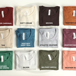K&ouml;nnte beinhalten: Eine Auswahl an gefalteten T-Shirts in verschiedenen Farben, darunter Heather Cardinal, Soft Cream, Brown und Heather Autumn. Weitere Farben sind Tan, Heather Deep Teal, Athletic Heather, Heather Maroon, Heather Grass Green, Silver, Military Green und Heather Sunset.