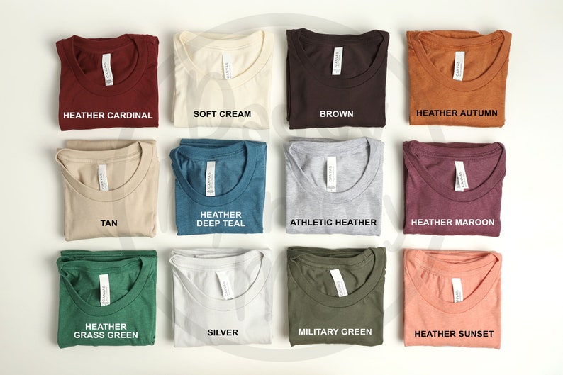 K&ouml;nnte beinhalten: Eine Auswahl an gefalteten T-Shirts in verschiedenen Farben, darunter Heather Cardinal, Soft Cream, Brown und Heather Autumn. Weitere Farben sind Tan, Heather Deep Teal, Athletic Heather, Heather Maroon, Heather Grass Green, Silver, Military Green und Heather Sunset.