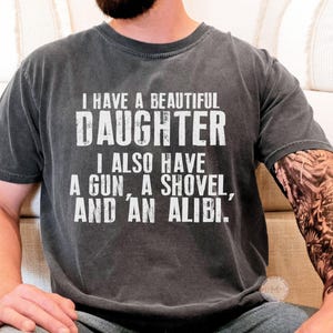 May include: A charcoal grey short-sleeved t-shirt featuring white distressed text that reads: I HAVE A BEAUTIFUL DAUGHTER I ALSO HAVE A GUN, A SHOVEL, AND AN ALIBI. The shirt has a crew neckline and a relaxed fit, showcasing a vintage-style wash.