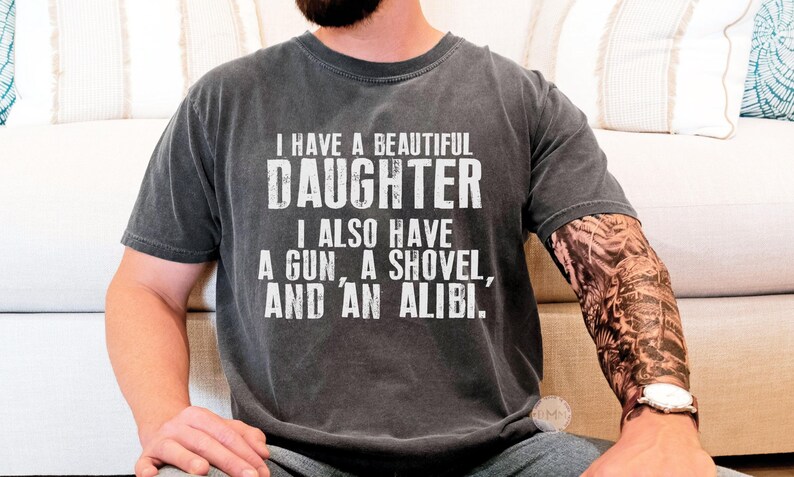 May include: A charcoal grey short-sleeved t-shirt featuring white distressed text that reads: I HAVE A BEAUTIFUL DAUGHTER I ALSO HAVE A GUN, A SHOVEL, AND AN ALIBI. The shirt has a crew neckline and a relaxed fit, showcasing a vintage-style wash.