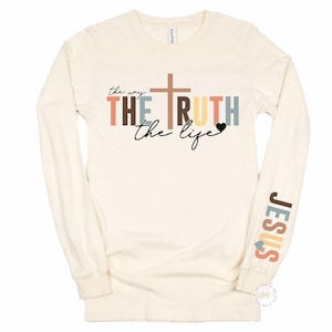 May include: Cream-colored long-sleeve shirt with the words "the way, the truth, the life" and a cross graphic. The word "Jesus" is printed on the sleeve in various colors. A heart graphic is also included.
