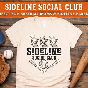 Op de afbeelding: Een cr&egrave;mekleurig t-shirt met een zwarte grafische print van drie klapstoelen, twee honkbalknuppels en een honkbal. De tekst SIDELINE SOCIAL CLUB staat in zwarte blokletters boven de afbeelding van de honkbal en knuppels.