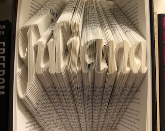 Puede incluir: Un libro con la palabra "Falicemal" doblada en las páginas. El libro está en un estante blanco con una cinta rosa atada alrededor.