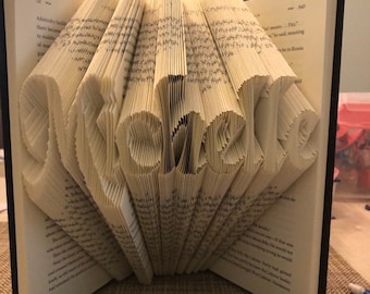 Puede incluir: Un libro con las páginas dobladas en forma de la palabra "remarkable". El libro está abierto y las páginas dobladas son visibles. El libro está sobre una superficie marrón.