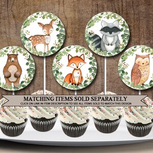 May include: Six cupcake toppers featuring woodland animals. The toppers are round and have a white background with green leaves around the edge. The animals include a deer, a raccoon, a bear, a fox, an owl, and a squirrel. The toppers are on white sticks and are ready to be placed on cupcakes.