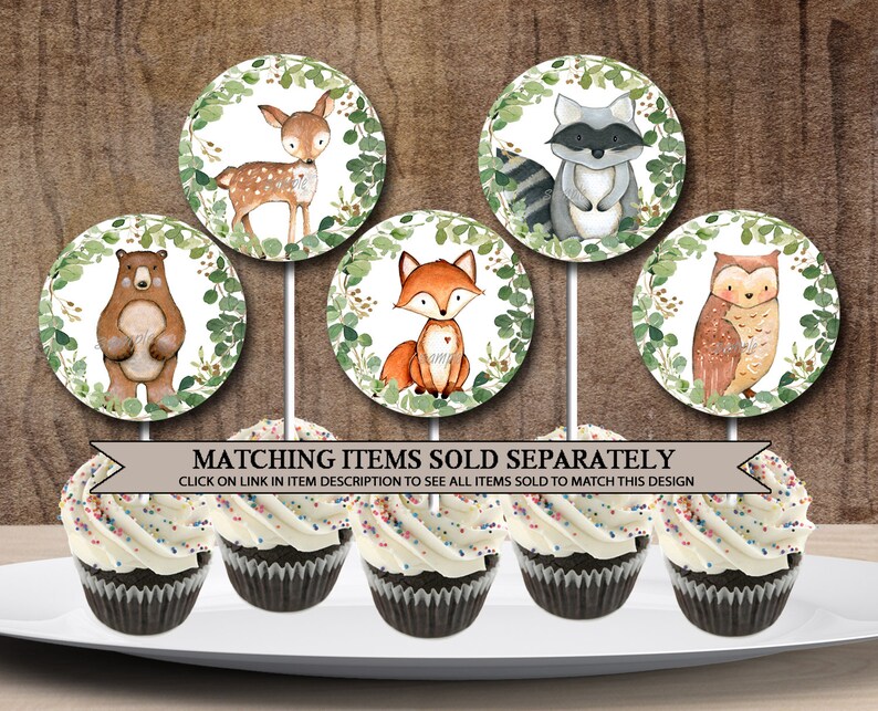 May include: Six cupcake toppers featuring woodland animals. The toppers are round and have a white background with green leaves around the edge. The animals include a deer, a raccoon, a bear, a fox, an owl, and a squirrel. The toppers are on white sticks and are ready to be placed on cupcakes.