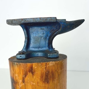 May include: A miniature blue anvil with a horn and flat top, set on a cylindrical wooden base. The anvil shows signs of wear and rust, while the wood has a natural grain and dark markings. A classic blacksmithing tool.